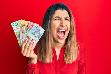 Canvas Print - Beautiful brunette woman holding australian dollars angry and mad screaming frustrated and furious, shouting with anger. rage and aggressive concept.