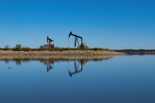 Pair Of Oil Pump Jacks On Rocky Peninsula In Glassy Calm Lake