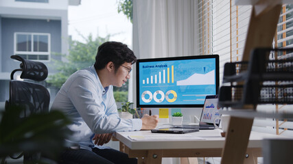 Candid of young attractive asian thai man busy work multiple screen computer or smart tablet on table desk at home in freelance data analyst, data science scientist for business.