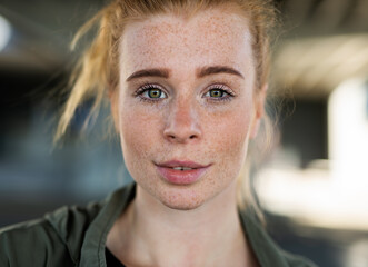 close-up portrait of young woman with red hair outdoors in town, looking at camera.