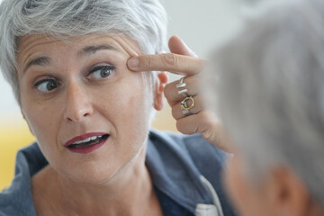 Wall Mural - Mature white-haired woman checking eye wrinkles in front of mirror