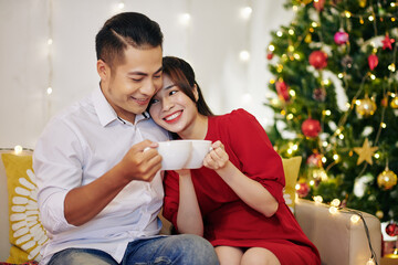 Sticker - Happy young Vietnamese couple enjoying hot chocolate when resting at home by Christmas tree