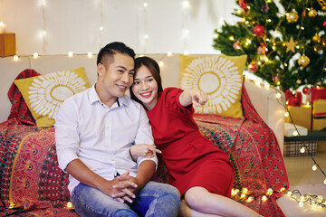 Poster - Beautiful smiling young Asian couple sitting on the floor in living room and watching tv