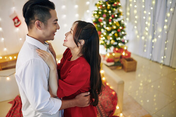 Poster - Happy young Asian boyfriend and girlfriend hugging and looking at each other when standing in room decorated for Christmas