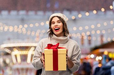 Sticker - winter holidays and people concept - happy smiling young woman in knitted hat and sweater holding gift box over christmas market lights on background