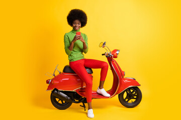 Wall Mural - Photo portrait of young brunette african american woman sitting on red scooter holding cellphone sending message wearing wearing green jumper isolated on vivid yellow colroed background