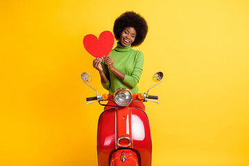 Sticker - Photo portrait of casual black skinned brunette woman on bike holding red heart cutout isolated on vivid yellow colored background