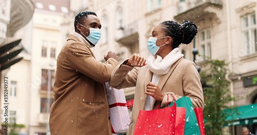 Obraz na płótnie Portrait of cheerful African American young man and woman in medical masks with xmas presents meeting on street after holiday shopping and kindly speaking while snowing. Friends greeting with elbows