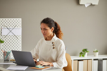 Portrait of modern young woman using laptop while working at desk in minimal office interior, copy space