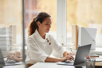 Portrait of successful businesswoman using laptop while sitting at desk by window and enjoying work in office, copy space