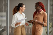 © Seventyfour - Waist up portrait of two successful young businesswomen smiling cheerfully looking at each other while standing in modern office interior, copy space