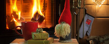 Mug Of Hot Chocolate Or Coffee, Gifts And Christmas Gnome On Vintage Wood Table In Front Of Fireplace As A Background.
