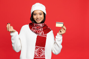 Wall Mural - smiling asian woman in hat and scarf holding credit card and showing money gesture isolated on red