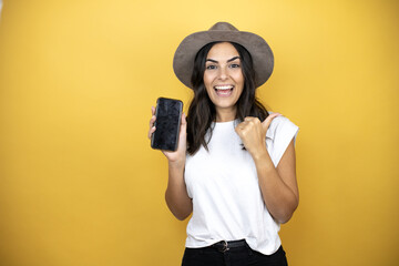 Beautiful woman wearing casual white t-shirt and a hat standing over yellow background smiling and pointing to the copy space