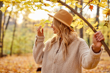 side view on blonde female in hat posing in autumn forest near the tree, she is looking away in contemplation