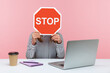© khosrork - Female office worker in striped shirt sitting at workplace hiding face behind red stop traffic sign avoiding conflicts, afraid of workplace bullying. Indoor studio shot isolated on pink background