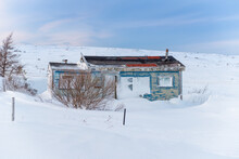 An Abandoned And Isolated Old Wooden And Small Farmhouse Sits In A Field Filled With White Fresh Snow. The Building Has A Small Window, A Door Drifted In With Snow And An Outhouse In The Back Yard. 