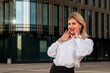 Stylish businesswoman in open air against background of business center. Woman in white blouse in business district. Portrait of pretty smiling female in style clothes at building. Copy space