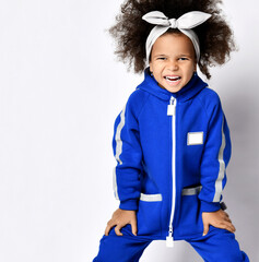 Positive cute curly african girl in comfortable blue sports jumpsuit and hair bow standing having fun over white wall background. Trendy children fashion, mixed race children, stylish outfit