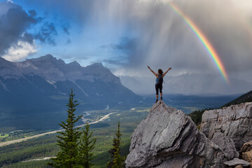 Wall Mural - Adventurous Caucasian Girl with open arms is on top of rocky mountain. Dramatic Sky with Rainbow. Taken from Mt Lady MacDonald, Canmore, Alberta, Canada.