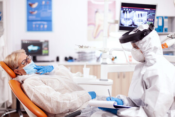 Dentist assitatnt questioning patient wearing protective equipment against coronavirus. Elderly woman in protective uniform during medical examination in dental clinic.