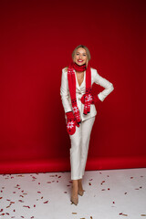 Full-length photo of a cheerful blonde lady wearing white suit and mittens with scarf while posing in red background