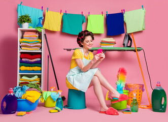 Sticker - Full body view of lovely cheerful maid sitting on bucket in messy room using device chatting app 5g isolated on pink pastel color background