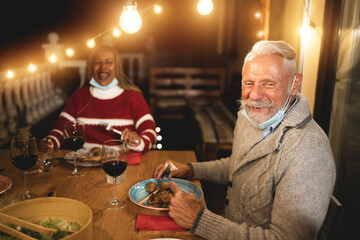 Wall Mural - Multiracial senior people celebrate christmas together with dinner outdoor while wearing safety mask for coronavirus outbreak - Soft focus on man face