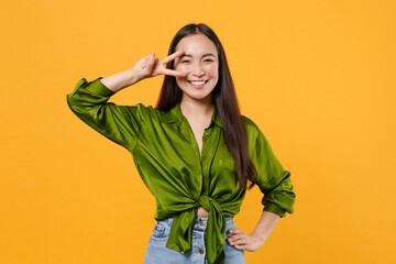 Wall Mural - Smiling cheerful funny attractive young brunette asian woman 20s wearing basic green shirt standing showing victory sign looking camera isolated on bright yellow colour background studio portrait.
