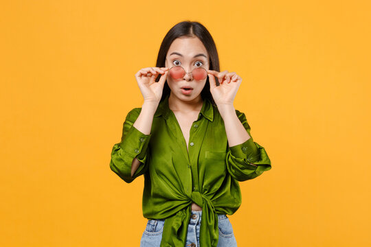 Shocked amazed young brunette asian woman 20s wearing basic casual green shirt eyeglasses standing keeping mouth open looking camera isolated on bright yellow colour wall background, studio portrait.