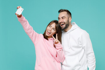 Wall Mural - Blinking funny young couple friends man woman in white pink casual hoodie doing selfie shot on mobile phone showing victory sign hugging isolated on blue turquoise colour background studio portrait.