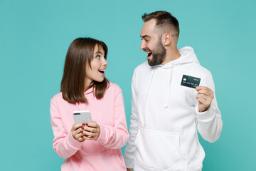 Wall Mural - Shocked amazed young couple two friends man woman 20s wearing white pink casual hoodie using mobile cell phone hold credit bank card isolated on blue turquoise colour wall background studio portrait.