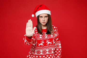 Displeased young brunette Santa woman 20s in sweater, Christmas hat showing stop gesture with palm isolated on bright red background, studio portrait. Happy New Year celebration merry holiday concept.