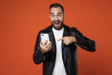 Amazed Young Bearded Man In Basic White T-shirt Black Leather Jacket Standing Pointing Index Finger On Mobile Cell Phone Typing Sms Message Isolated On Bright Orange Colour Background Studio Portrait.