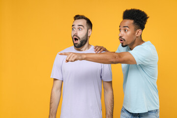 Wall Mural - Shocked young two friends european african american men 20s in violet blue casual t-shirts pointing index finger aside keeping mouth open isolated on bright yellow colour background studio portrait.