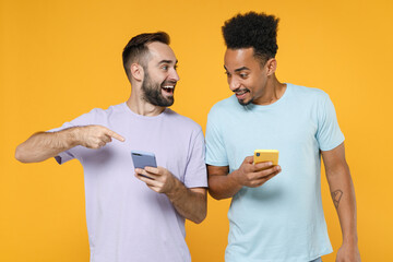 Excited young two friends european african american men 20s in violet blue casual t-shirts pointing index finger on mobile cell phone typing sms message isolated on yellow background studio portrait.