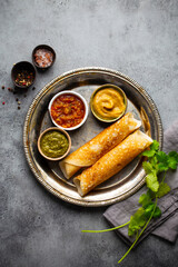 Wall Mural - Traditional Indian rice pancakes Dosa with different dips chutney and seasonings on rustic metal plate on stone background table. Quick meal or vegetarian snack of South India, top view