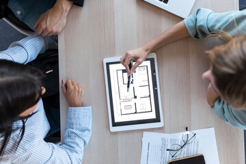 Beautiful real-estate agent showing house plans on electronic tablet while talking to the couple about buying the house in the office.