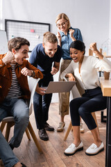 excited multicultural businesspeople screaming and showing success gesture while looking at laptop