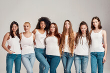 Group of beautiful diverse young women wearing white shirt and denim jeans looking at camera while posing together isolated over grey background