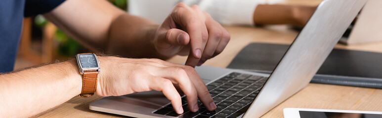 Wall Mural - Cropped view of man with watch typing on laptop, while sitting at desk with blurred african american woman on background, banner