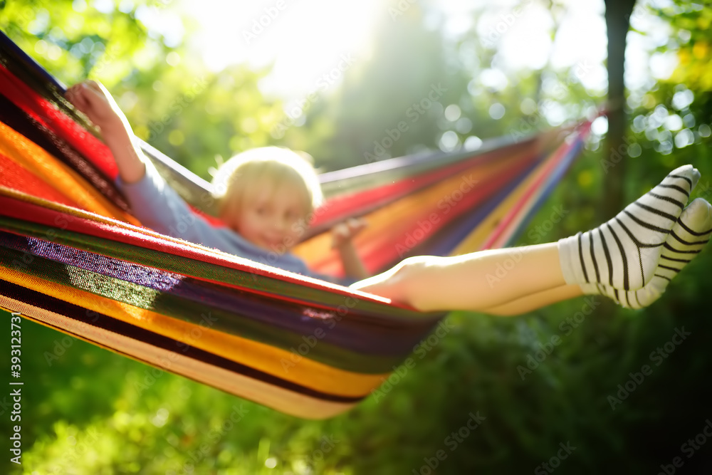 Cute little blond caucasian boy having fun with multicolored hammock in backyard or outdoor playground. Summer active leisure for kids. Child swinging and relaxing in hammock. - obrazy, fototapety, plakaty 