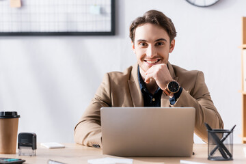 smiling office worker looking at camera while sitting near laptop at desk in office on blurred backg