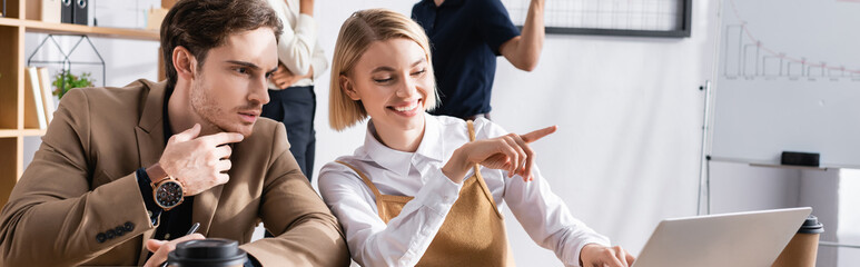 Focused man looking at laptop, while sitting near happy woman pointing with finger, with colleagues on background, banner