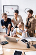 © LIGHTFIELD STUDIOS - Blonde woman sitting at table with skeptical, multicultural office workers standing behind, gesturing and looking at laptop at workplace