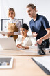 © LIGHTFIELD STUDIOS - Skeptical office workers standing near african american woman and looking at laptop on table with blurred stationery on foreground