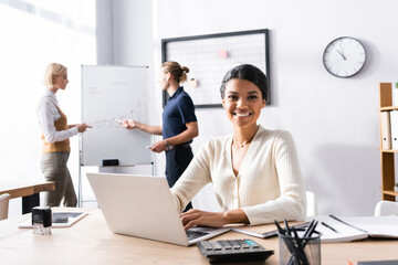 Wall Mural - Smiling african american woman looking at camera, while typing on laptop with blurred office workers on background