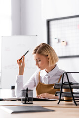 Focused blonde woman with pen looking at notepad while sitting at desk in office on blurred background