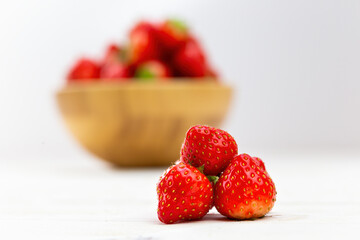 Little pile of strawberries with wooden bowl in background. Fresh red small berries on heap on white table. Bowl full of fresh healthy fruit blurred in backdrop.