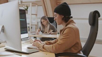 Pan shot of young Caucasian male coder wearing hoody, hat and round-shaped glasses sitting at workplace, developing program code on computer and then looking at camera